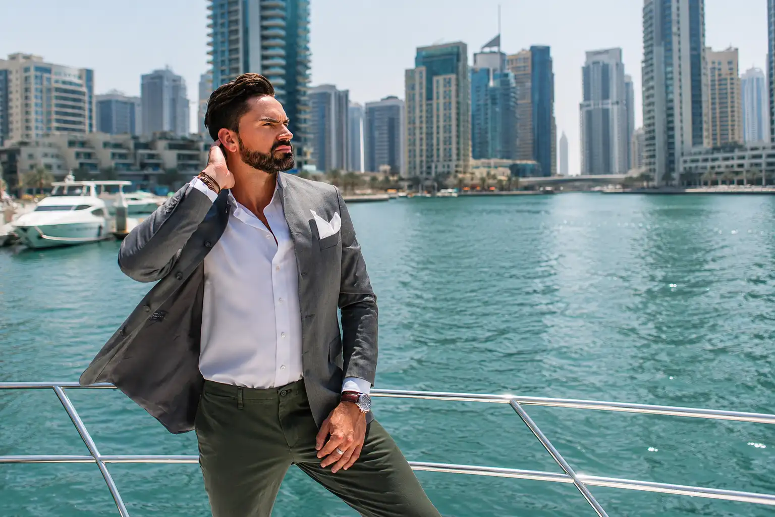 Professional Dubai brand ambassador standing aboard a luxury yacht, adjusting his cuff while wearing a light blue blazer and sunglasses, with Dubai's waterfront skyline and turquoise waters creating a sophisticated backdrop.