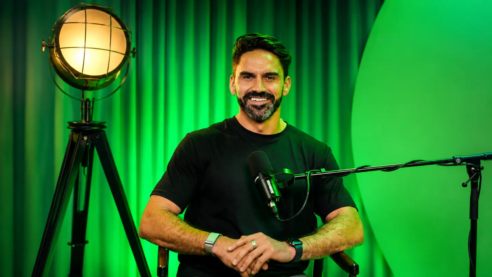 Professional event MC Darryl Reeves in a modern podcast studio, smiling behind a Shure microphone with cinematic green lighting, showcasing expertise in live event hosting, public speaking, and corporate entertainment.