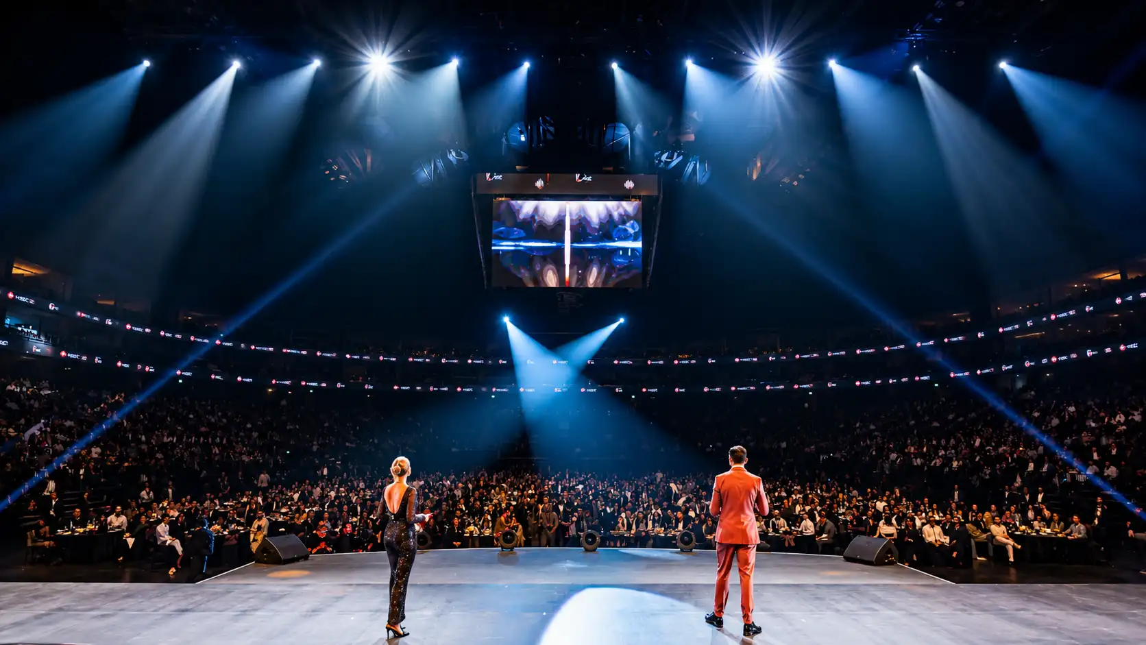 Wide view of a luxury corporate awards event with professional MCs on stage addressing a packed arena audience under dramatic lighting, showcasing large scale live event hosting, audience engagement, and premium event production.