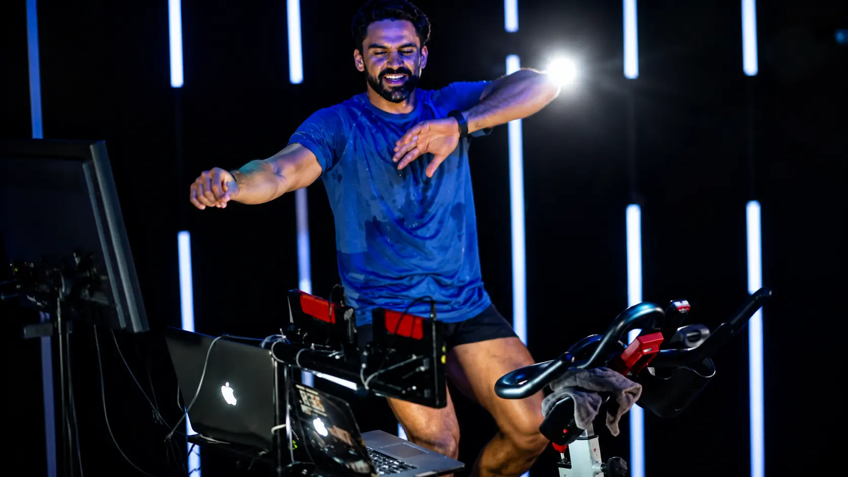 Indoor cycling instructor leading a high-energy spin class on a stationary bike, reaching forward while riding in a modern fitness studio with LED light bars, workout equipment, and a laptop controlling the music and class experience.