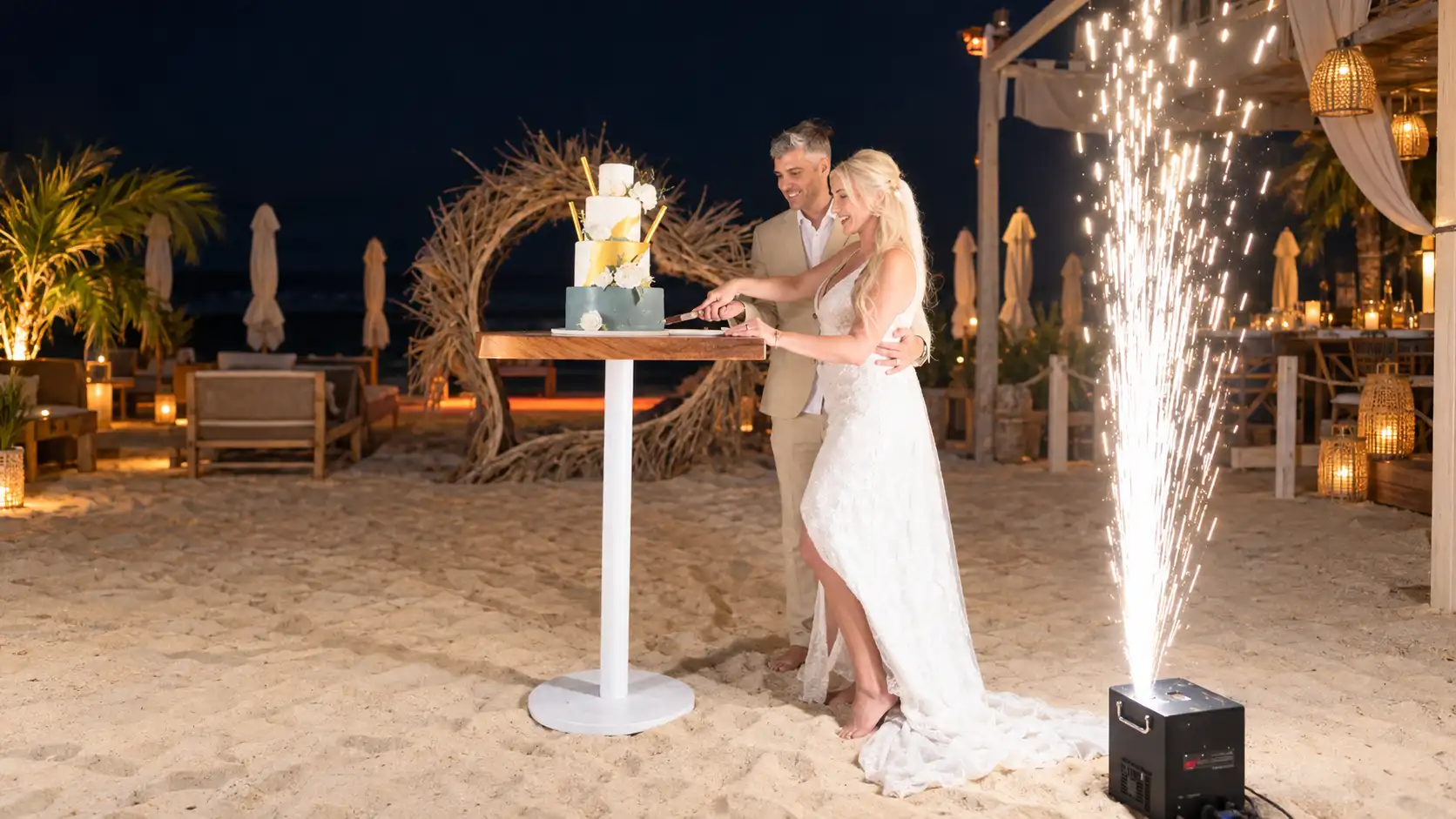 Victoria and Wes cutting their wedding cake during a nighttime beach celebration as a cold spark fountain lights up beside them.