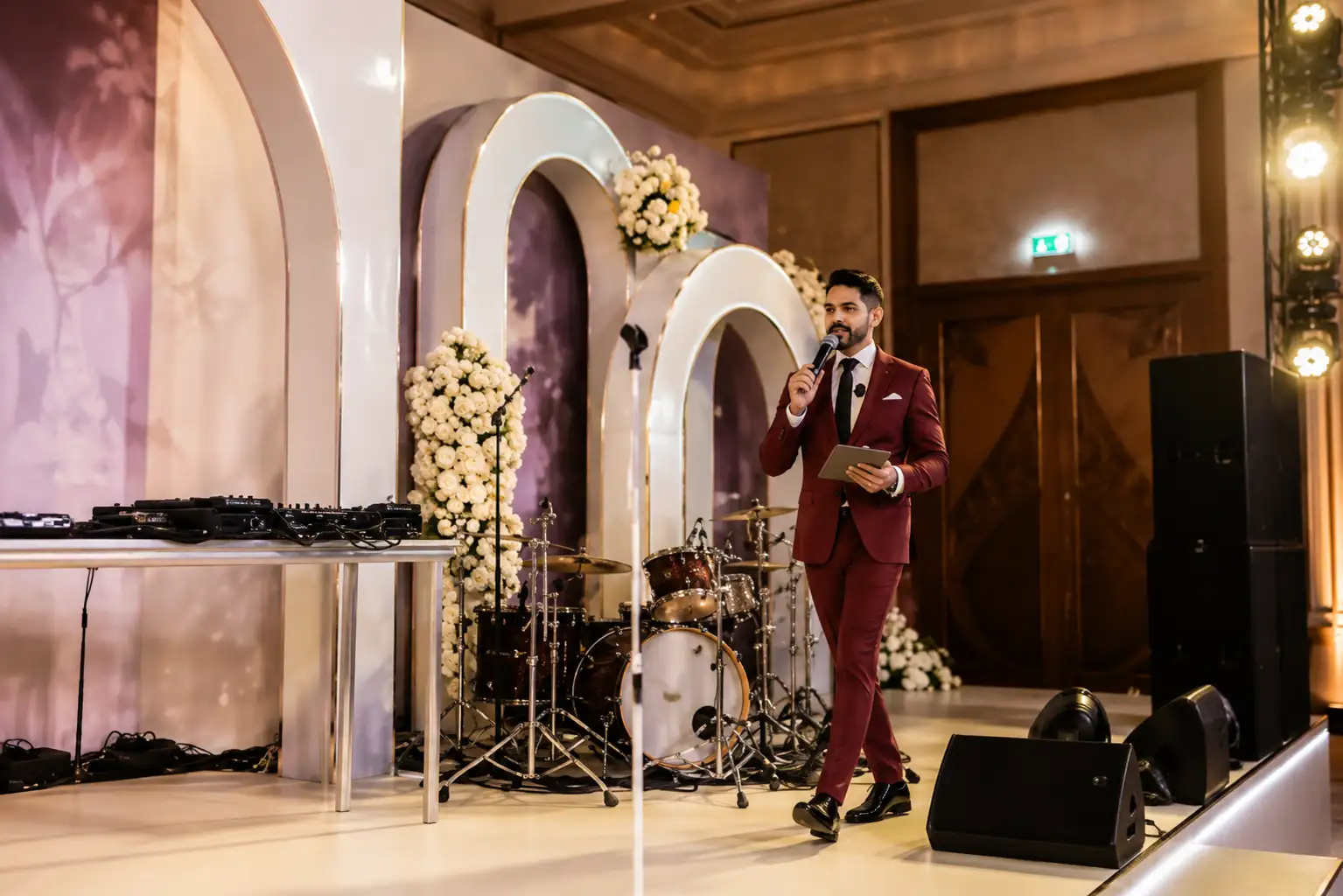 Professional wedding MC in a burgundy suit walking across a beautifully decorated reception stage in Dubai, hosting the celebration with a microphone and tablet beside the DJ setup and live band.